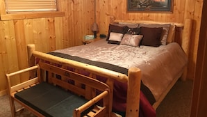 Een slaapkamer in een houten chalet, met een bed, kussens, een nachtkastje, een lamp en een ingelijste foto aan de muur.