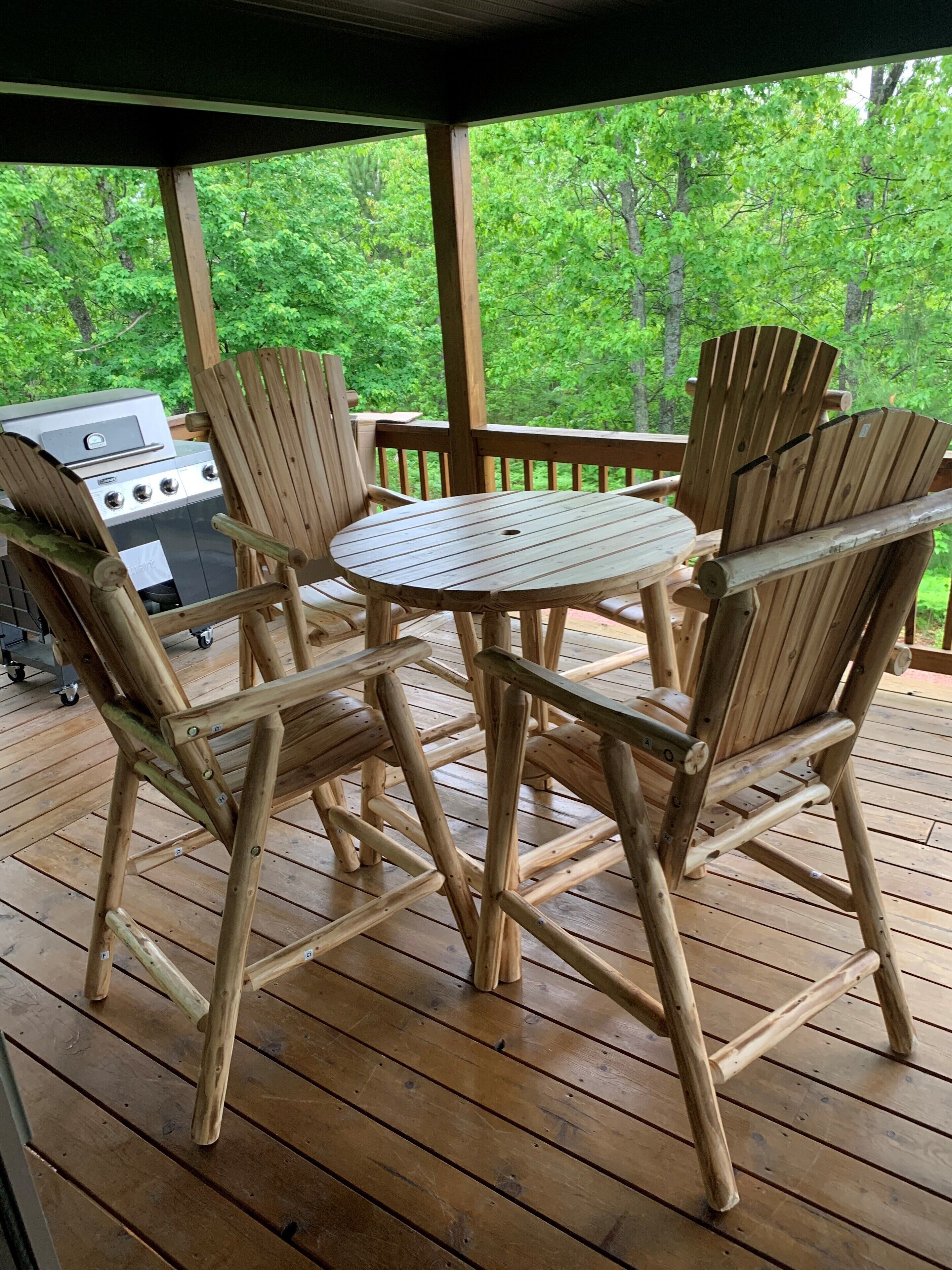 A wooden deck with a round table and four chairs, a grill, and a view of green trees.