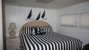 Ein Schlafzimmer mit gestreifter Bettdecke, einem Kopfteil, einem Nachttisch mit Lampe und einer Wanddekoration mit Segelbooten.