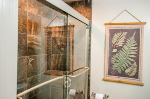 A bathroom with a glass shower enclosure, a wooden-framed picture of ferns, and a marble-tiled wall.