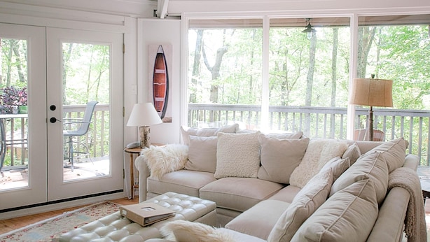 A cozy living room with a beige sofa, a small table, and a chair. There is a large window with a view of trees and a balcony.