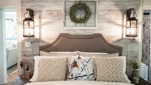 A bedroom with a wooden headboard, two wall-mounted lanterns, a decorative wreath, and patterned pillows.