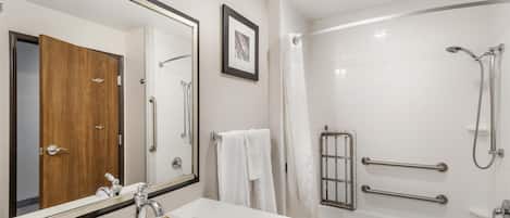A modern bathroom with a large mirror, a wooden door, a white sink with toiletries, a bathtub, and a shower with grab bars.