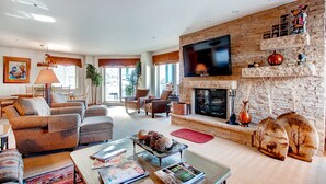 A spacious living room with a stone fireplace, comfortable seating, a flat-screen TV, and a large window providing natural light.