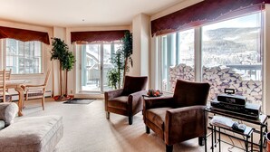 A spacious living room with a large window offering a mountain view, two brown armchairs, a wooden dining table with chairs, and a small side table with a radio.
