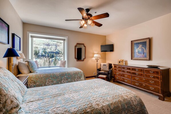A bedroom with two beds, a ceiling fan, a dresser, a mirror, and a window with a view of a mountainous landscape.