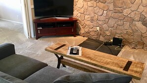 Een woonkamer met een stenen muur, een flatscreen televisie op een houten dressoir, een salontafel met een afstandsbediening en een smartphone, en grijze banken.