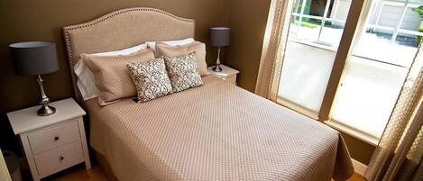 Una cama bien hecha con una colcha beige texturizada, dos almohadas a juego y un cabecero. Hay dos mesitas de noche con lámparas, una a cada lado de la cama. La habitación tiene suelo de madera y una ventana con cortinas.