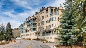 A multi-story building with balconies, a curved driveway, and surrounding trees. - Centennial 2 Bedroom Condo Unit no. 7 (Avon)