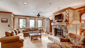 A spacious living room with a fireplace, a ceiling fan, and large windows.