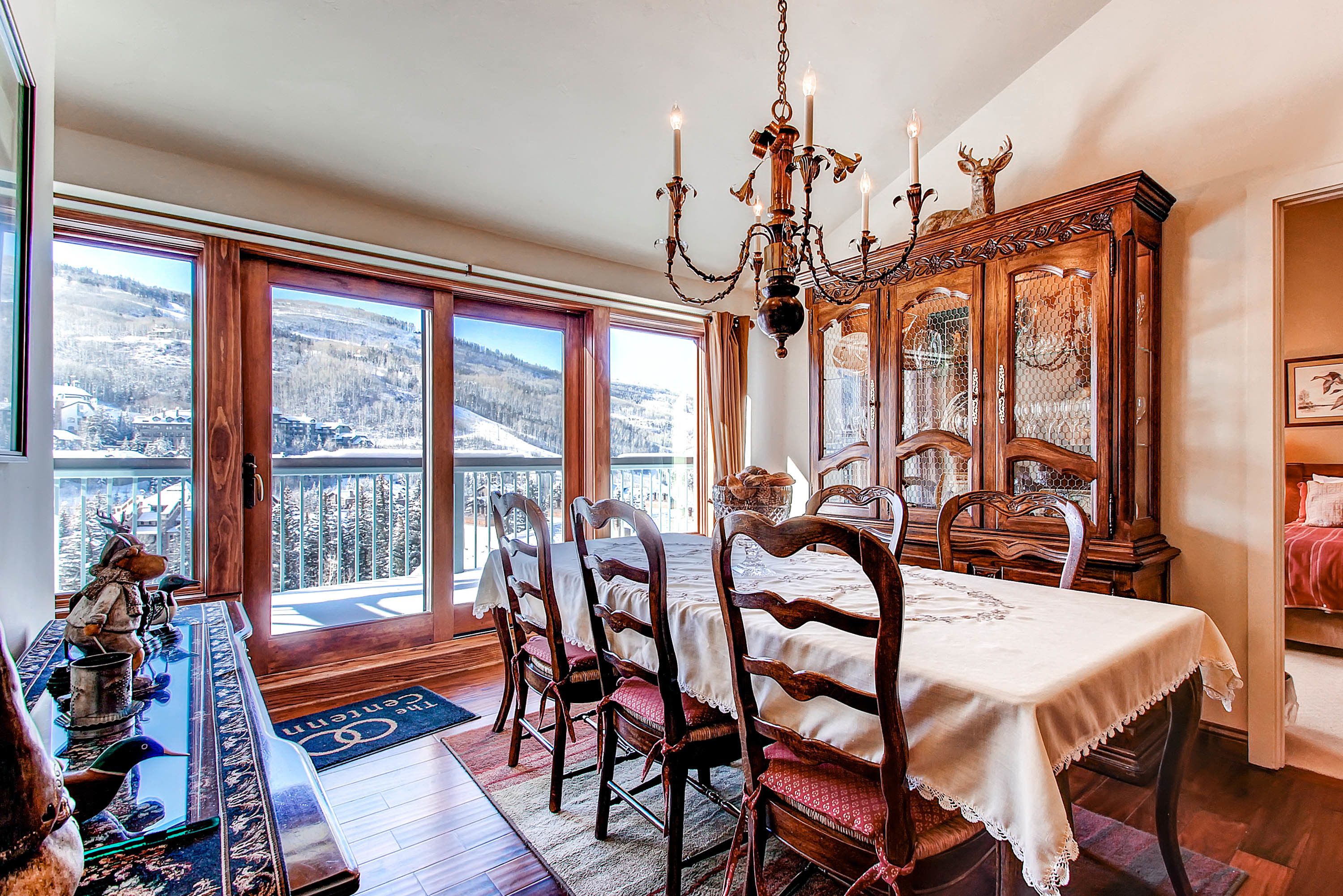 A dining area with a table set for four, surrounded by wooden chairs, a chandelier, and a view of a snowy landscape through large windows.