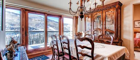 A dining area with a table set for four, surrounded by wooden chairs, a chandelier, and a view of a snowy landscape through large windows.