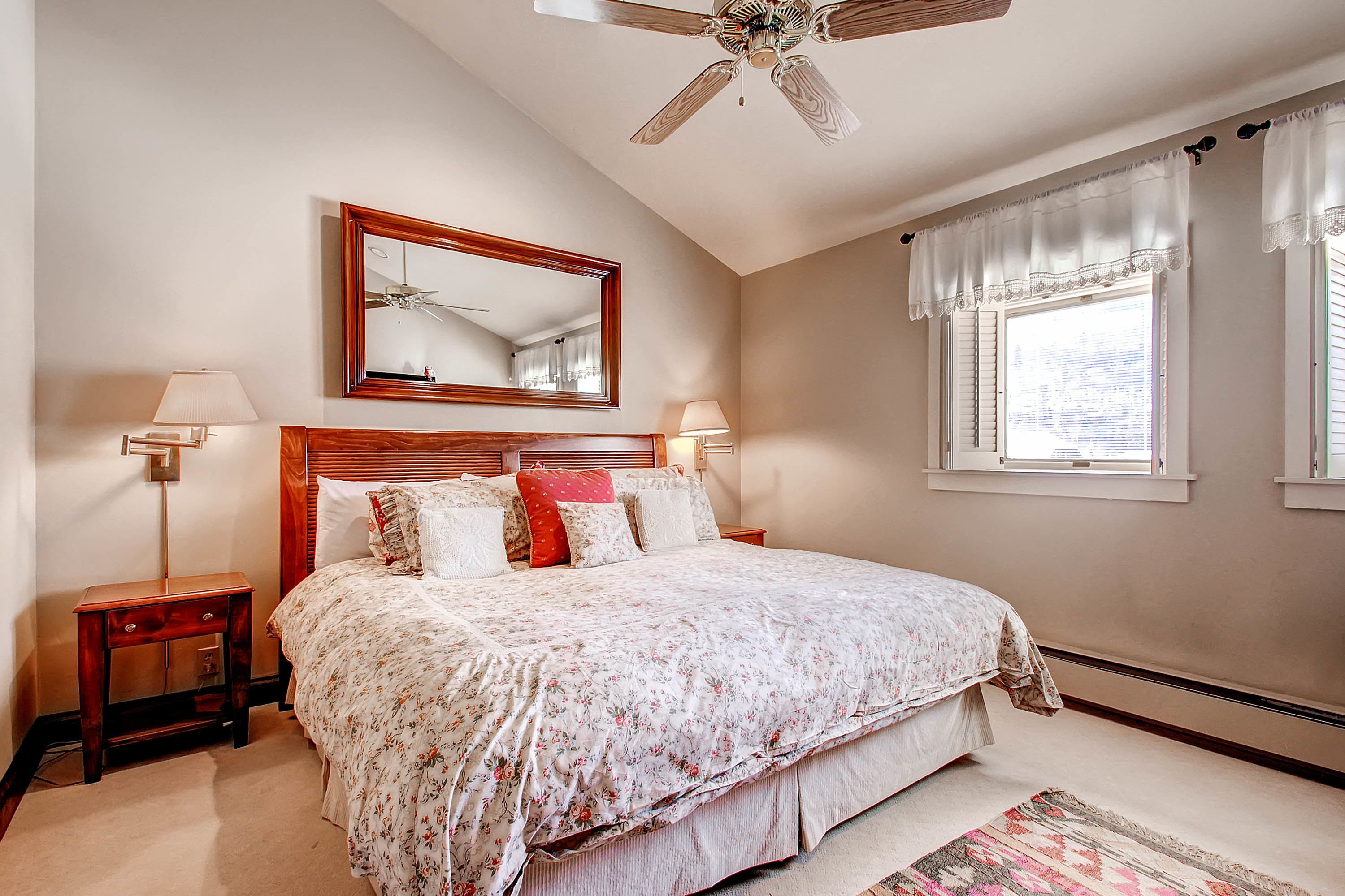 A bedroom with a large bed, two bedside tables, a ceiling fan, a window with curtains, and a mirror on the wall.