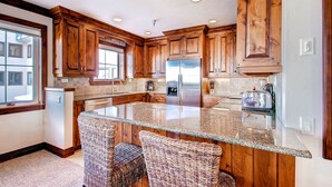 A kitchen with wooden cabinets, a granite countertop, and a built-in island with two wicker chairs. - Centennial 2 Bedroom Condo Unit no. 3 (Avon)