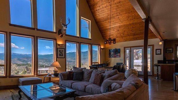 Ein geräumiges Wohnzimmer mit einer großen Eckcouch, einem Glastisch und einem Kamin. Mehrere große Fenster bieten Blick auf die Berge.