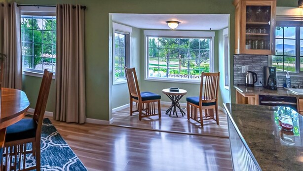 Een eetgedeelte met een houten tafel en stoelen, een kleine ronde tafel met een stoel, en uitzicht op een groen landschap door grote ramen.