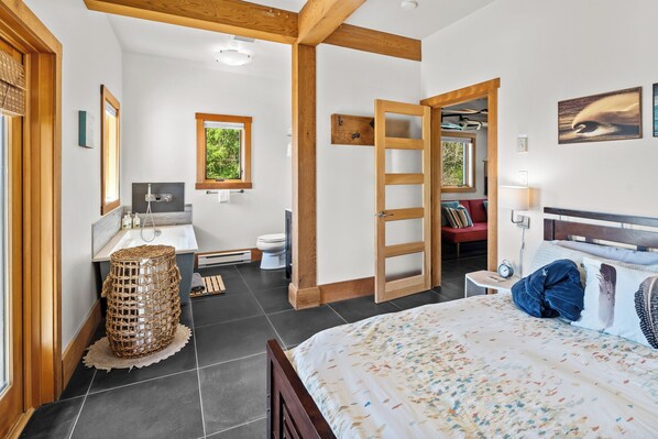 A bedroom with a bed, a bedside table, a nightstand, a window, a door, and a bathroom with a sink and toilet.