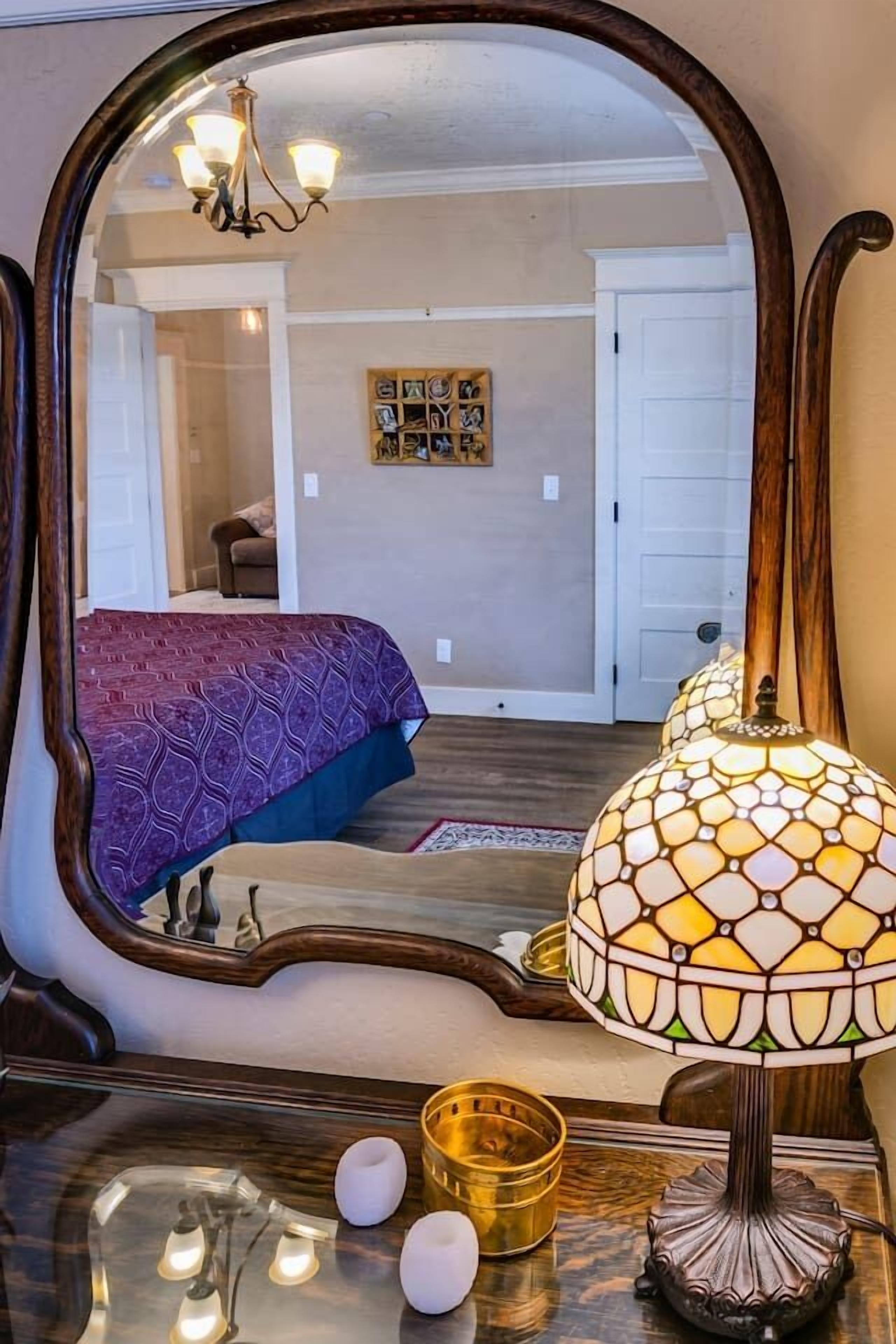 A bedroom with a large mirror, a patterned bedspread, a decorative lamp, and a small table with a candle and a bowl.