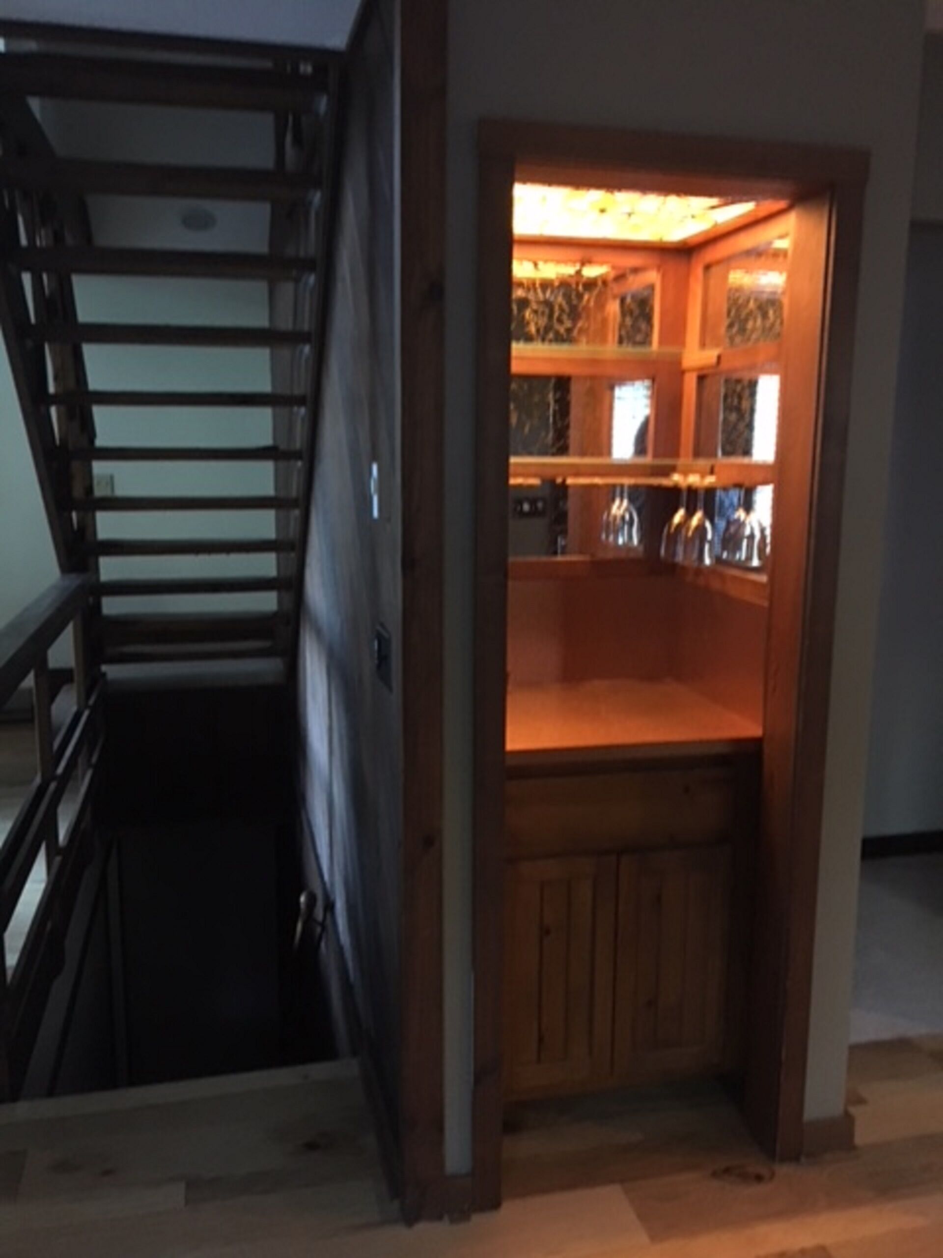 Una escalera de madera que desciende a un nivel inferior, un pequeño armario de madera con puerta de vidrio y un estante con diversos objetos.
