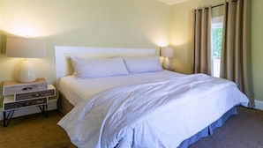 Una cama bien hecha con edredón blanco y almohadas, una mesita de noche con lámpara y una ventana con cortinas.