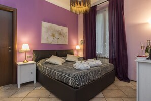 A bedroom with a bed, two bedside lamps, a painting, a window with curtains, and a nightstand with a bottle and glasses.
