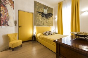 Una habitación con cama, una silla, una mesita de noche, un cuadro en la pared y una ventana con cortinas.