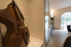 Une pièce comprenant un violoncelle, un miroir, une chaise noire et donnant sur une cuisine et un coin repas.