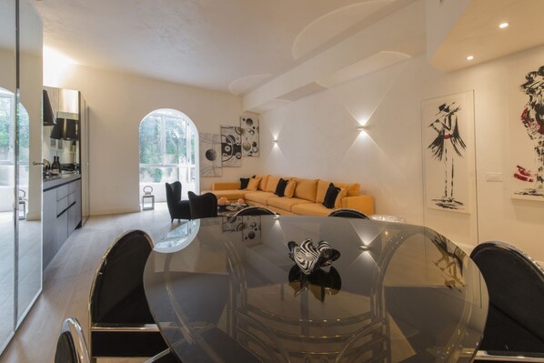 A modern dining area with a glass table, black chairs, a sofa with orange cushions, and abstract wall art.