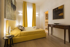 A room with a bed, a bedside table, a lamp, a painting, and a window with curtains.