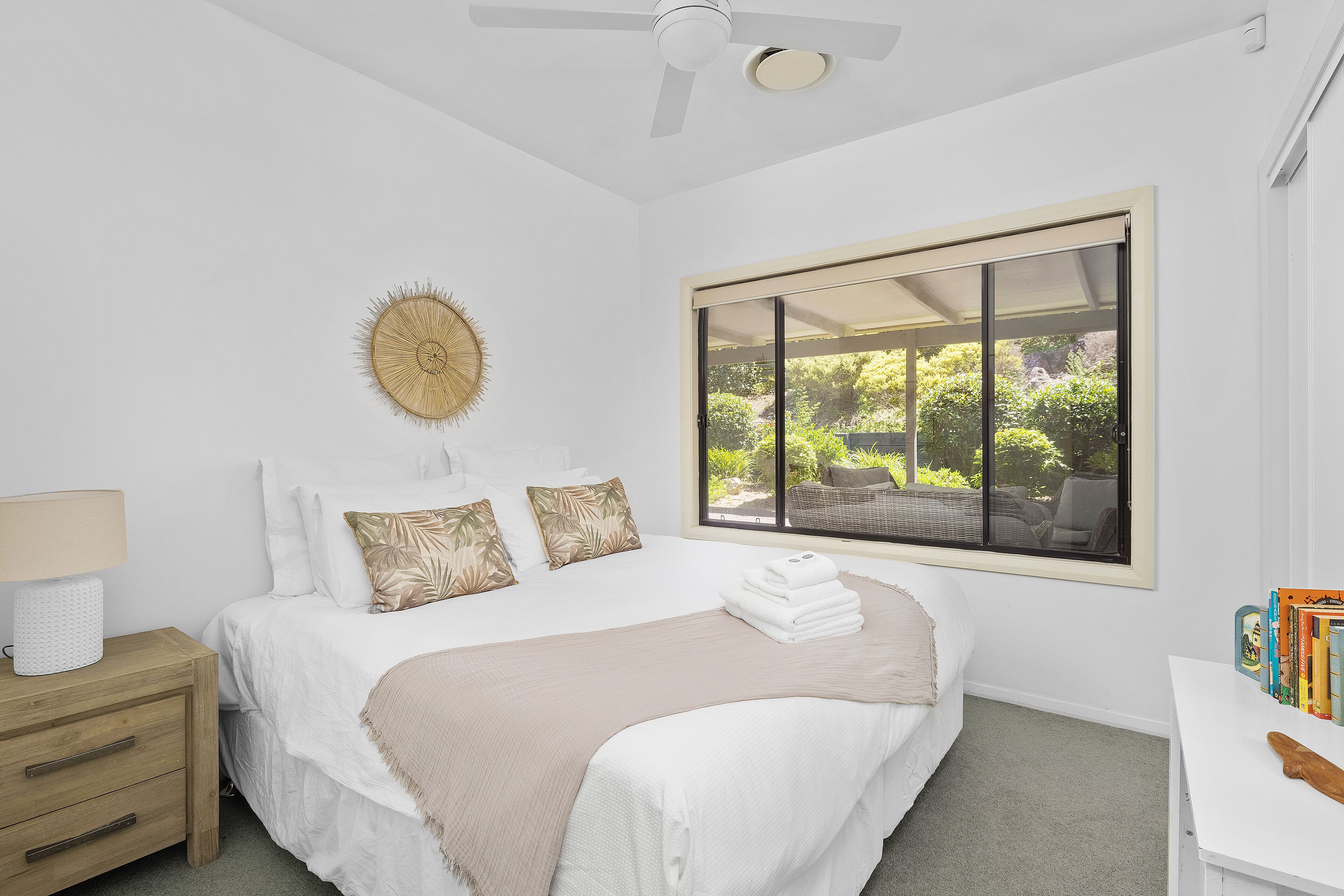 A bedroom with a bed, a nightstand, a window with a view of greenery, and a ceiling fan.