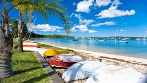 A beach with colorful boats lined up on the shore, a clear blue sky, and a calm sea with sailboats. - Wanda Point House - pure tranquillity and walk to beach (Salamander Bay)