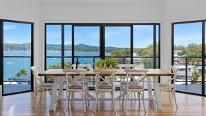 A dining area with a wooden table and white chairs, large windows with a view of a body of water and hills. - Wanda Point House - pure tranquillity and walk to beach (Salamander Bay)
