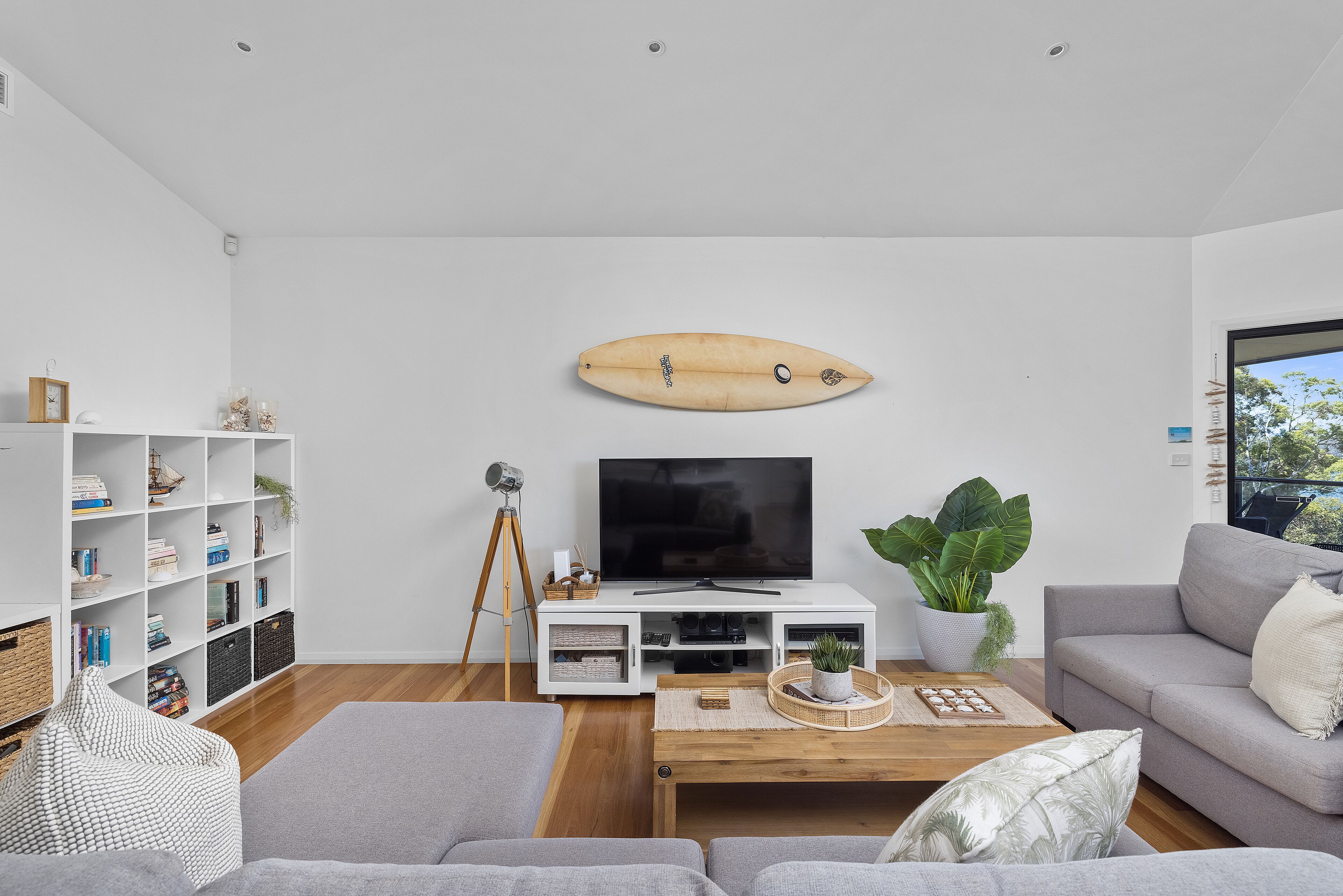 Una sala de estar moderna con un televisor de pantalla plana, una decoración con una tabla de surf, un sofá gris, una mesita de centro y una estantería.