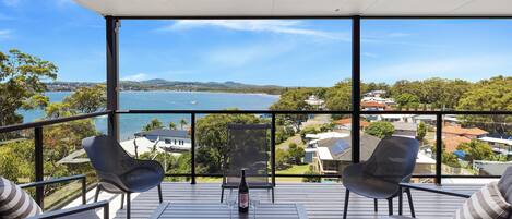 A balcony with a table, two chairs, and a bottle of wine, overlooking a scenic bay with boats and houses.