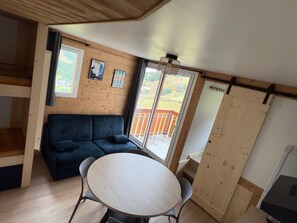 A room with a wooden ceiling, a dark blue sofa, a round table, and a view of a balcony with a railing. - Apartment in Les Menuires, 4 pers, ski-in/ski-out (Les Menuires)