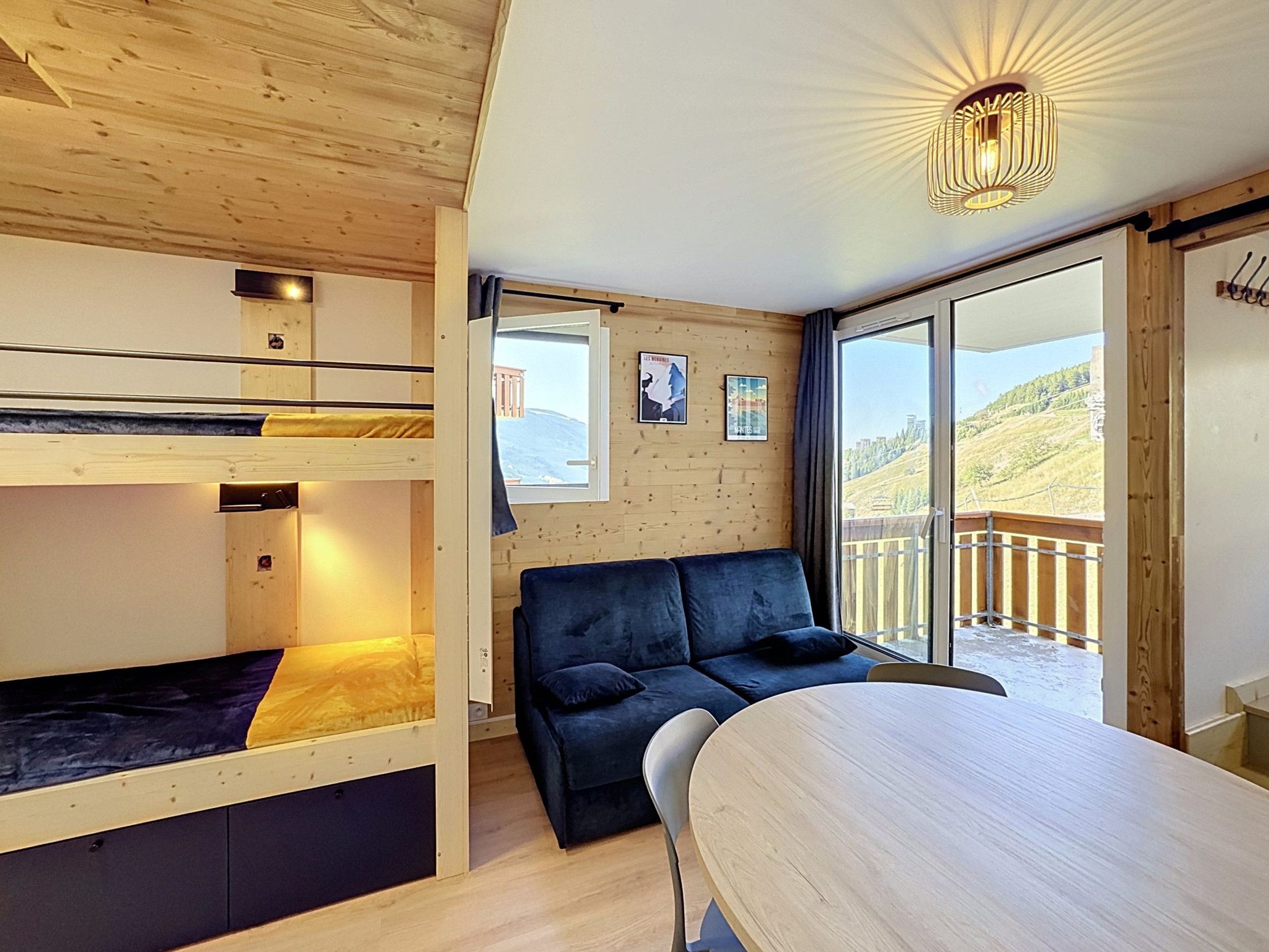 A room with bunk beds, a sofa, a dining table, and a balcony with a view of a hilly landscape.