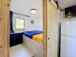 A compact room with a bed, a small table, a window, and a white refrigerator.