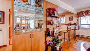 Una cocina con armarios de madera, una vitrina de vidrio con utensilios de cocina, un taburete de metal y una ventana con vistas a árboles.