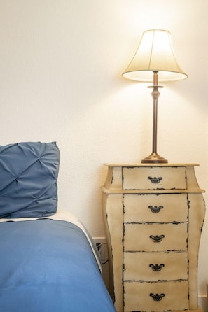 Een bed met een blauwe spreideken en een wit kussen, een nachtkastje met een lamp, en een dressoir met vier lades.
