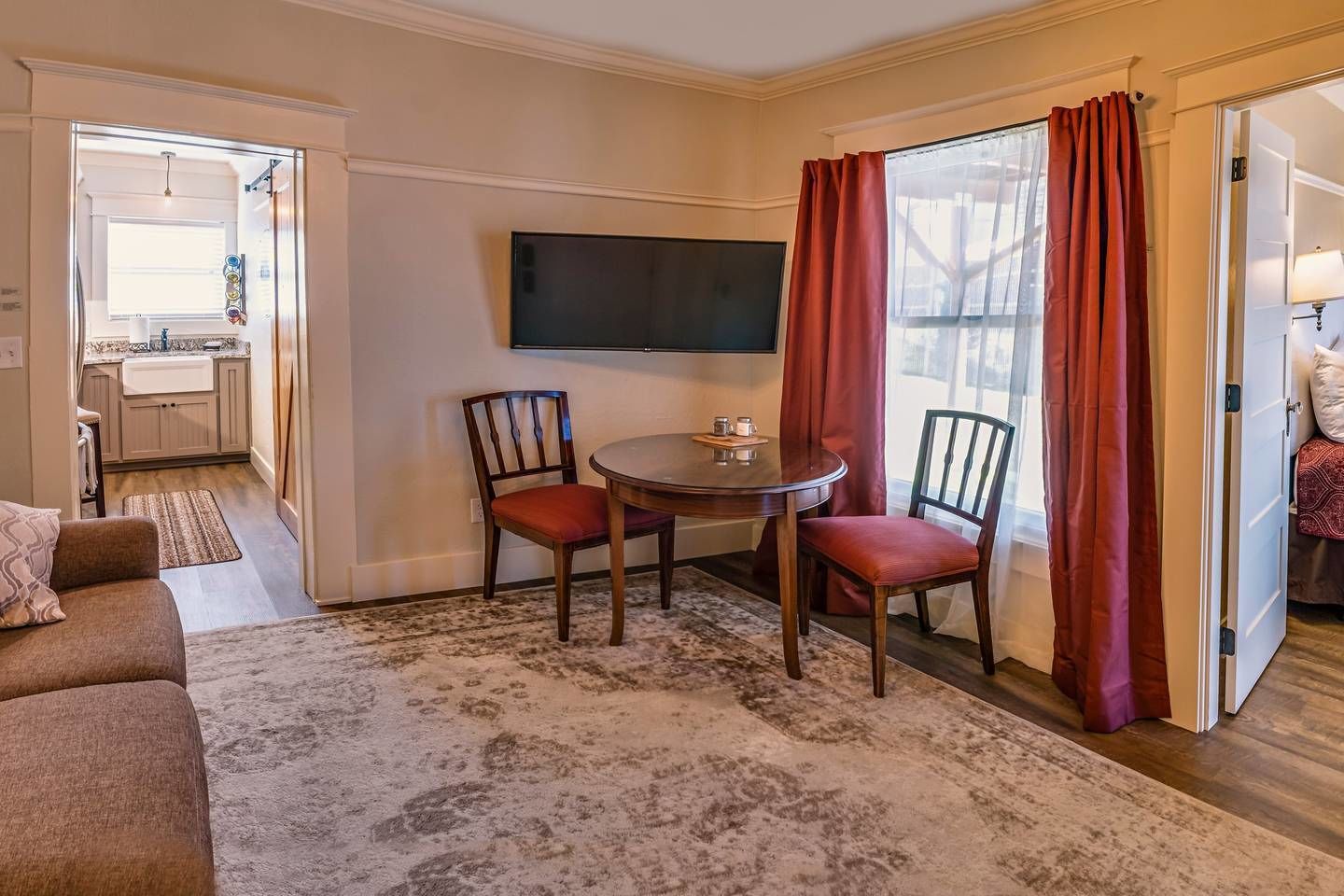 A cozy living room with a sofa, a coffee table, and two chairs. A flat-screen TV is mounted on the wall, and there is a window with red curtains.