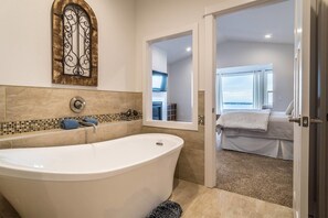 Un baño moderno con una bañera independiente, un espejo decorativo y vista a un dormitorio con cama y ventana.
