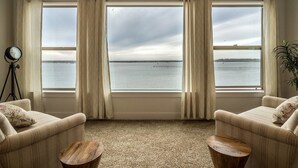 Ein Zimmer mit zwei Sofas, die zu einem großen Fenster mit Blick auf ein Gewässer zeigen, einer Uhr auf einem Stativ und einem kleinen Tisch.