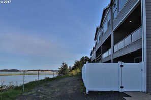 Un edificio moderno con fachada blanca, zona de estacionamiento cubierta y vista a un cuerpo de agua y terreno distante.