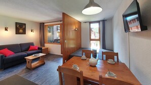 A cozy living room with a sofa, coffee table, and dining table with chairs. There is a television mounted on the wall and a window with curtains.