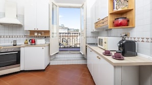 Een compacte keuken met witte kastjes, een ingebouwde oven, een magnetron en een broodrooster. Er is een klein raam dat uitkomt op een balkon met uitzicht op gebouwen.
