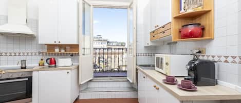 A compact kitchen with white cabinets, a built-in oven, a microwave, and a toaster. There is a small window leading to a balcony with a view of buildings.