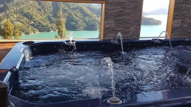 A hot tub with water jets, set against a backdrop of a mountainous landscape through a large window. - Luxury Self Contained Unit,Wide Views (Portage)