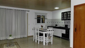 Cocina compacta con armarios blancos, una mesa de comedor con cuatro sillas, un ventanal con persianas y una puerta de madera.