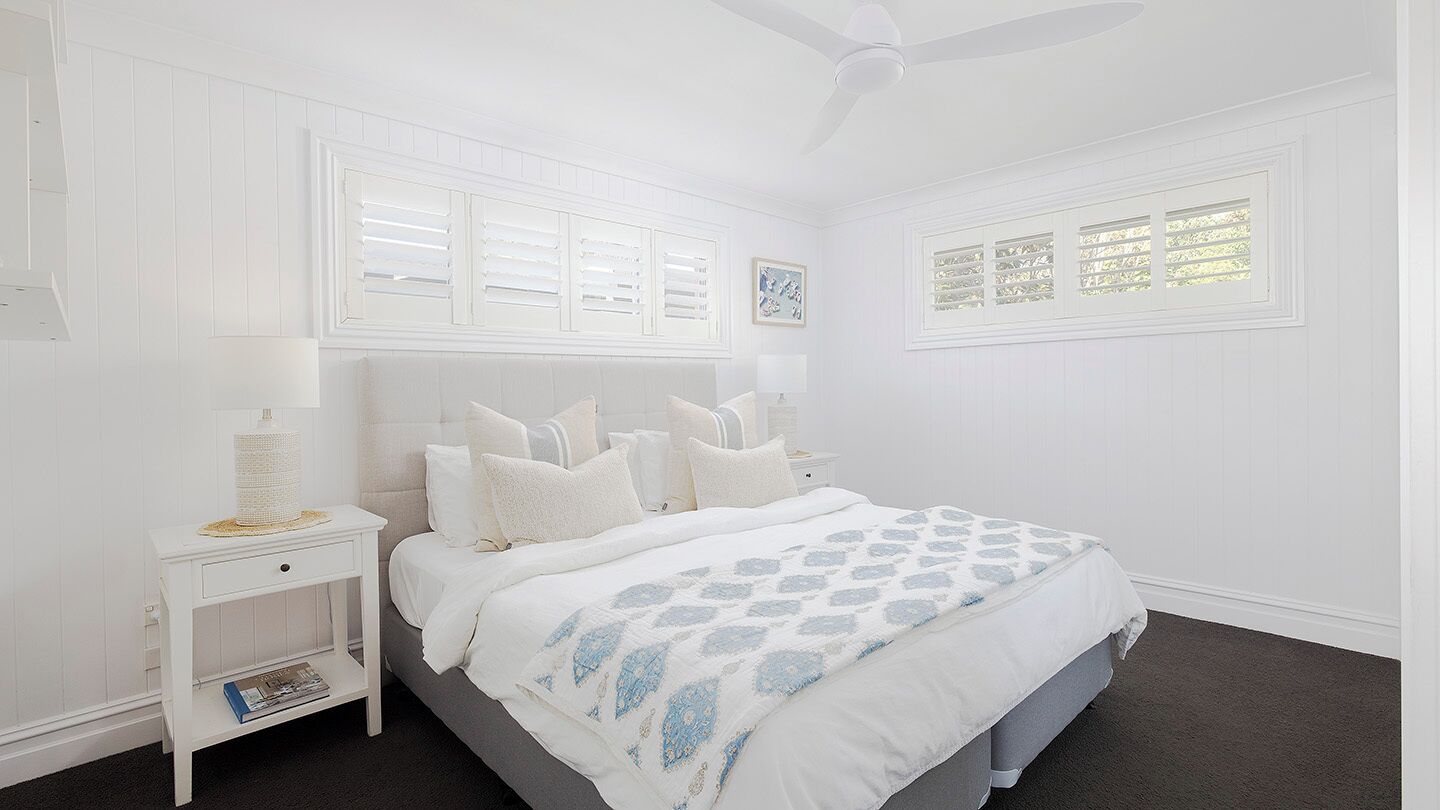 A bedroom with a bed, white nightstands, a ceiling fan, and a window with shutters.