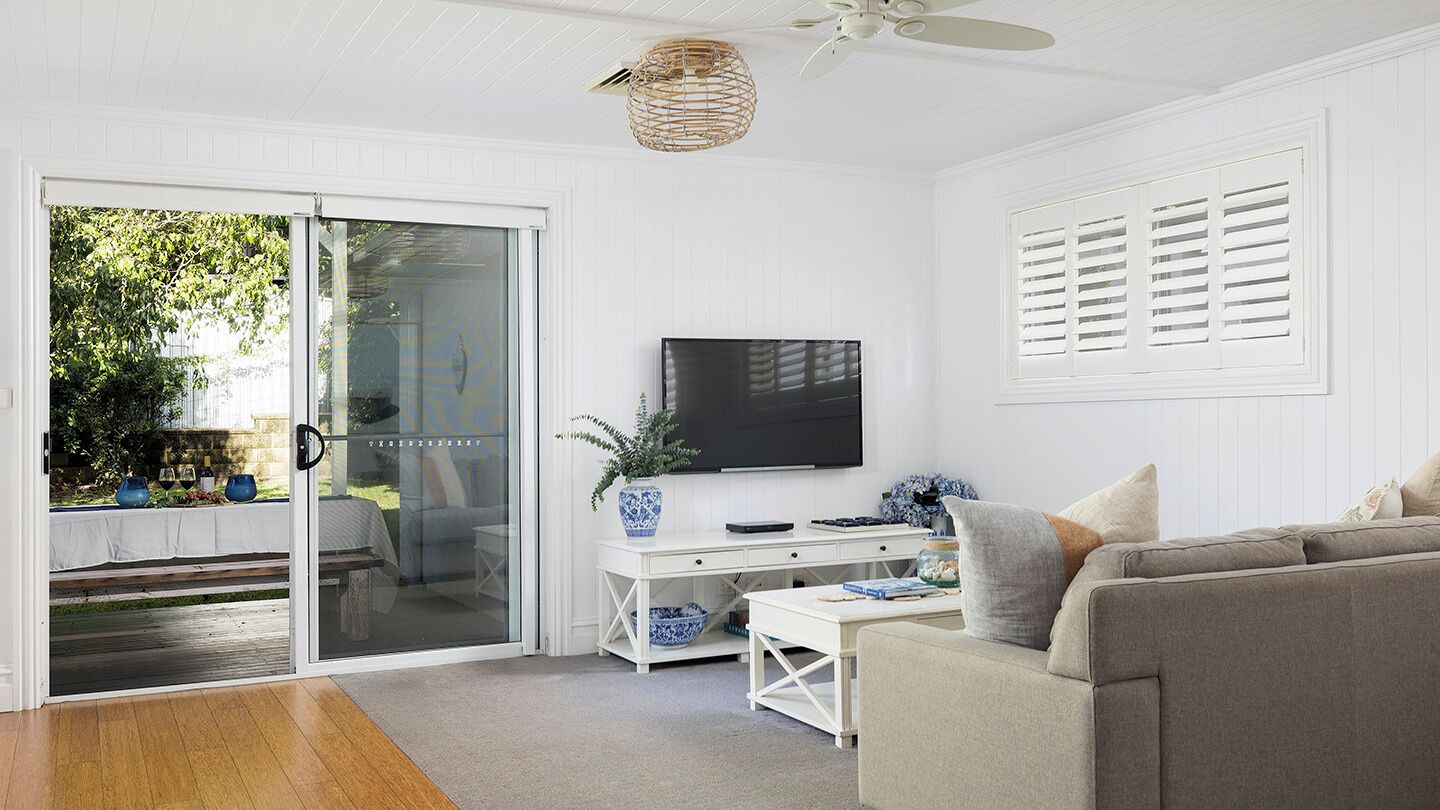 A living room with a sliding glass door leading to a patio, a flat-screen TV mounted on the wall, a white desk with a vase, and a grey sofa.